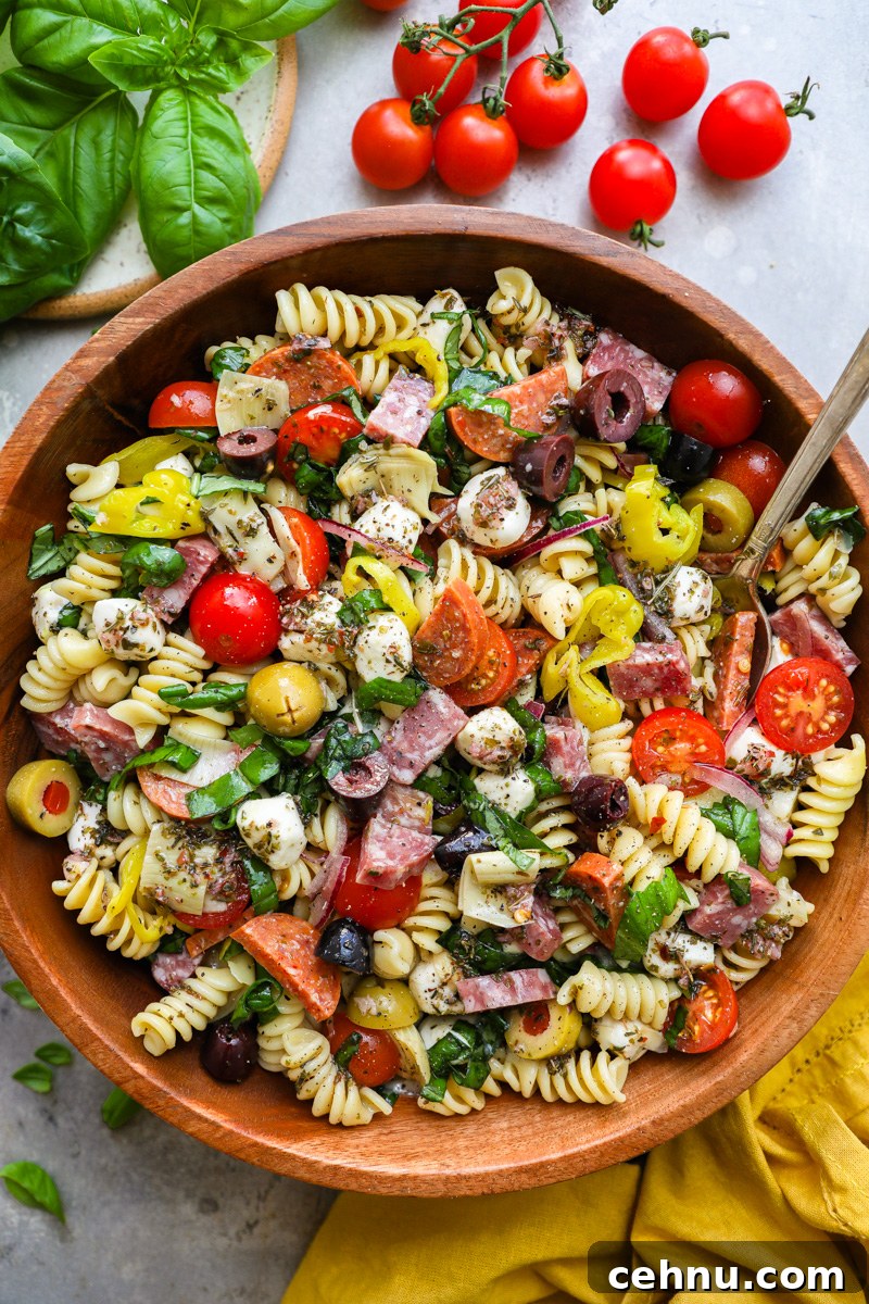 A large wooden serving bowl filled with antipasto pasta salad sitting next to fresh basil and cherry tomatoes