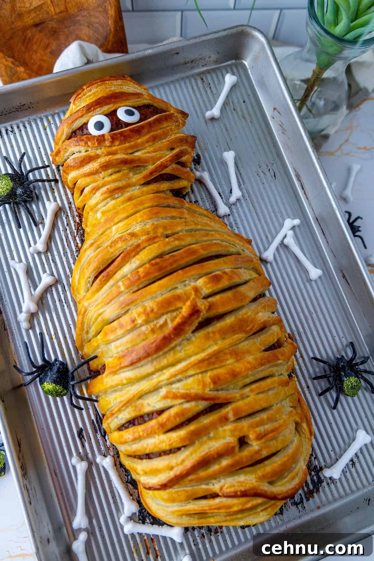 Halloween Mummy Meatloaf Recipe 2 A tray features a mummy-shaped meatloaf with googly eyes, surrounded by decorative bones and spiders.
