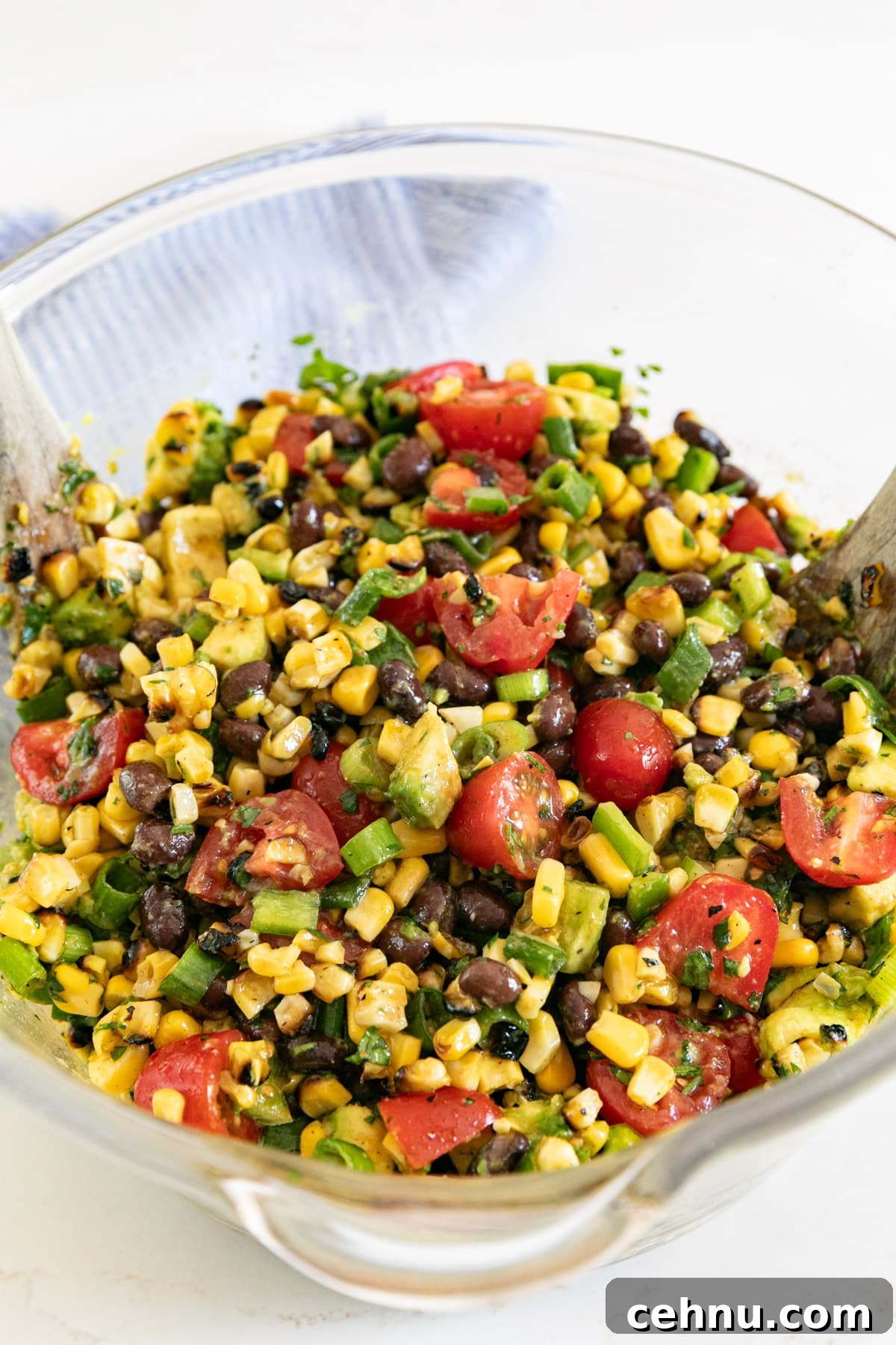 A large glass bowl of cajun corn salad with wooden salad servers in it.
