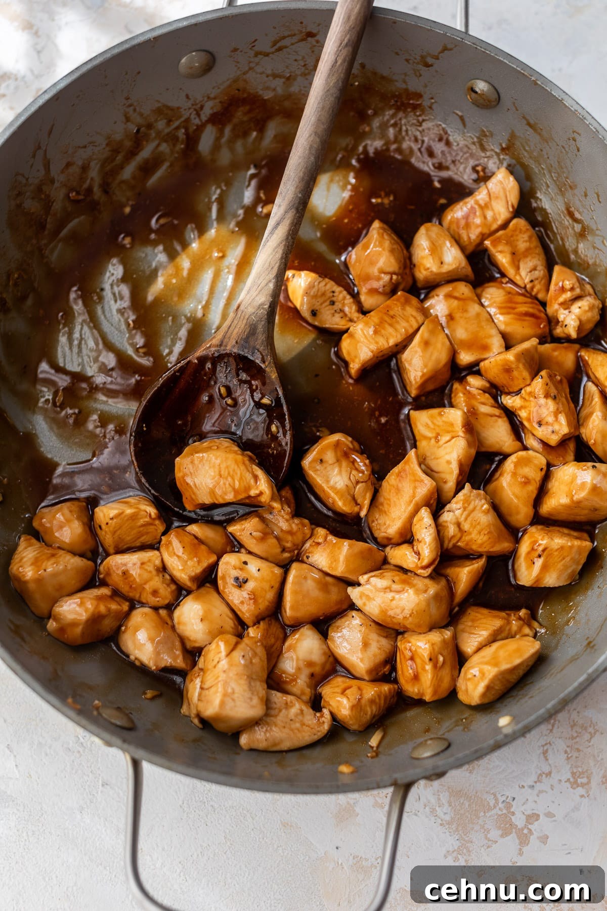 overhead view of a wooden spoon stirring teriyaki sauce-covered cooked chicken bites in a large skillet.