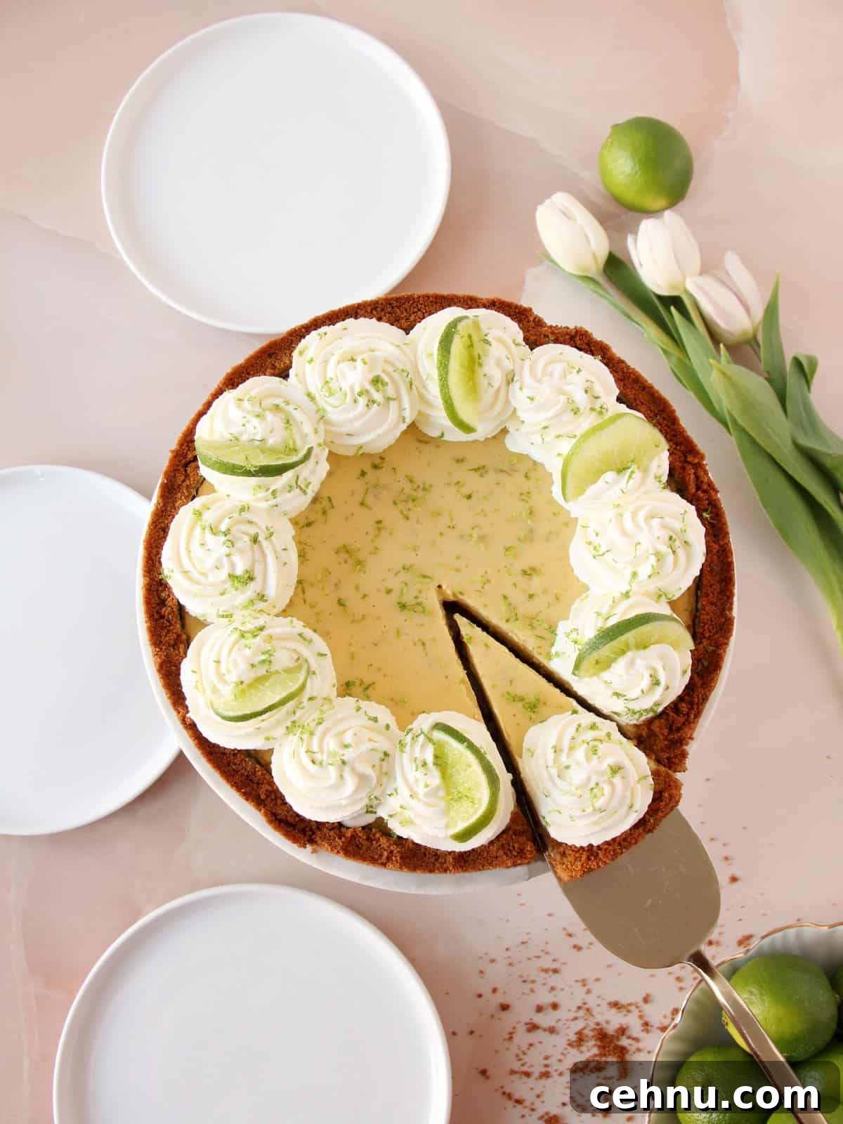 Keylime pie on cake stand, decorated with swirls of whipped cream, wedges of lime and lime zest. Three white round plates ready to be served. White tulips and limes as decoration on pink marble background.