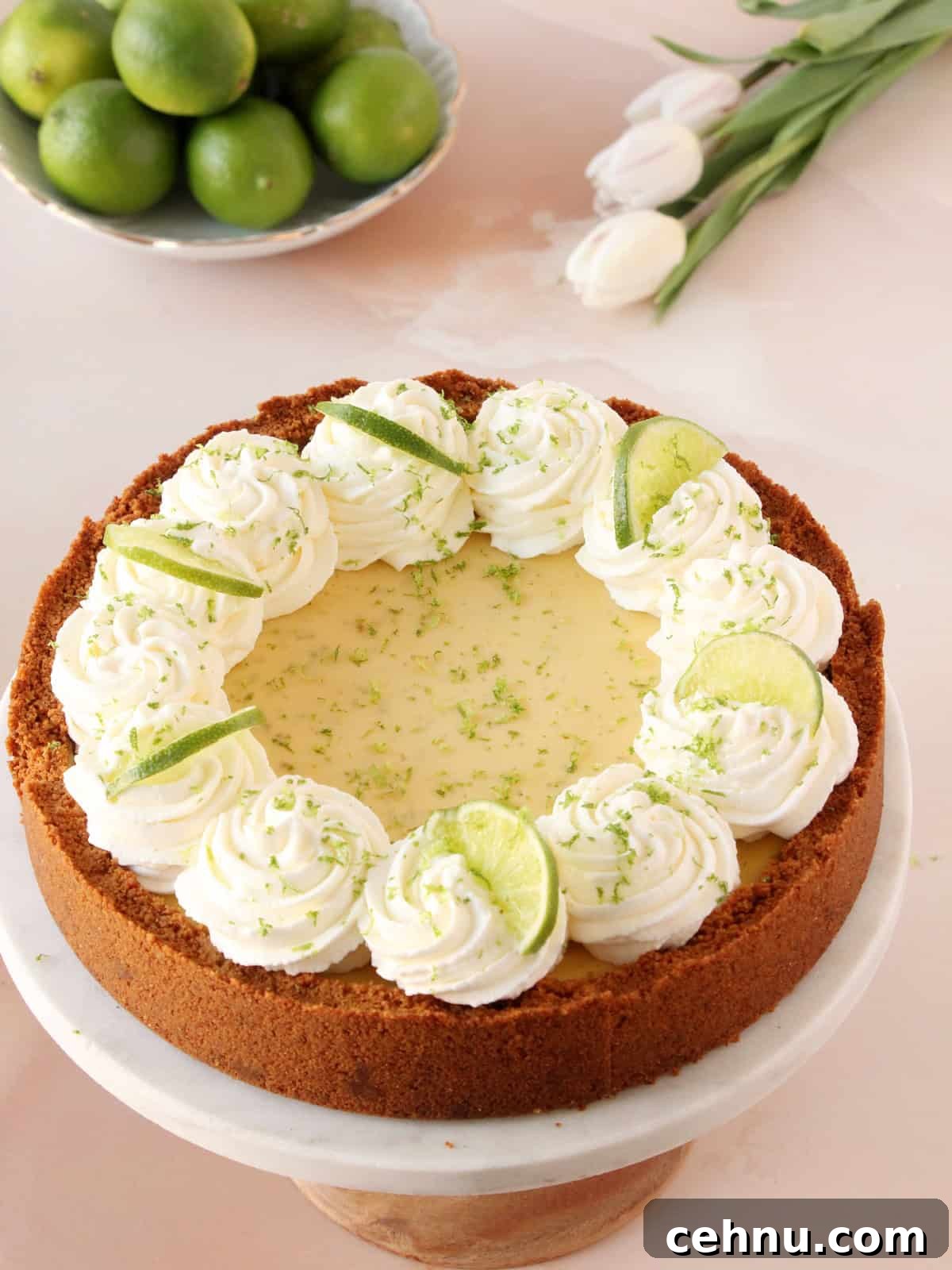 Key lime pie placed on cake stand with limes and white tulips in background.