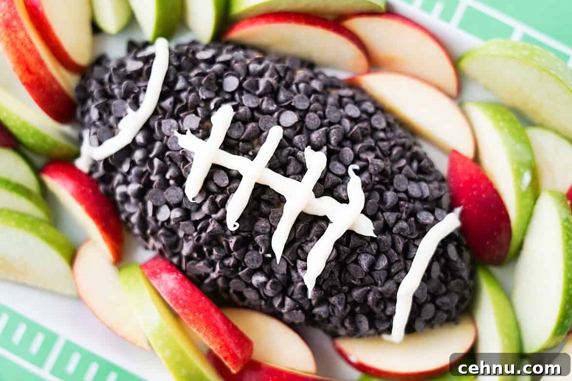 apple toffee dip shaped into a football, coated in chocolate chips and white icing for lines in the football on serving tray surrounded with apple slices