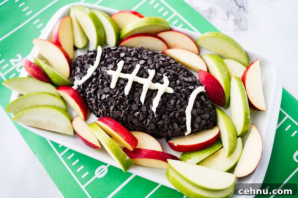 45 degree shot of apple toffee dip on white serving platter with dip shaped as a football, coated in chocolate chips and white icing for laces on the football, served with slices of apples