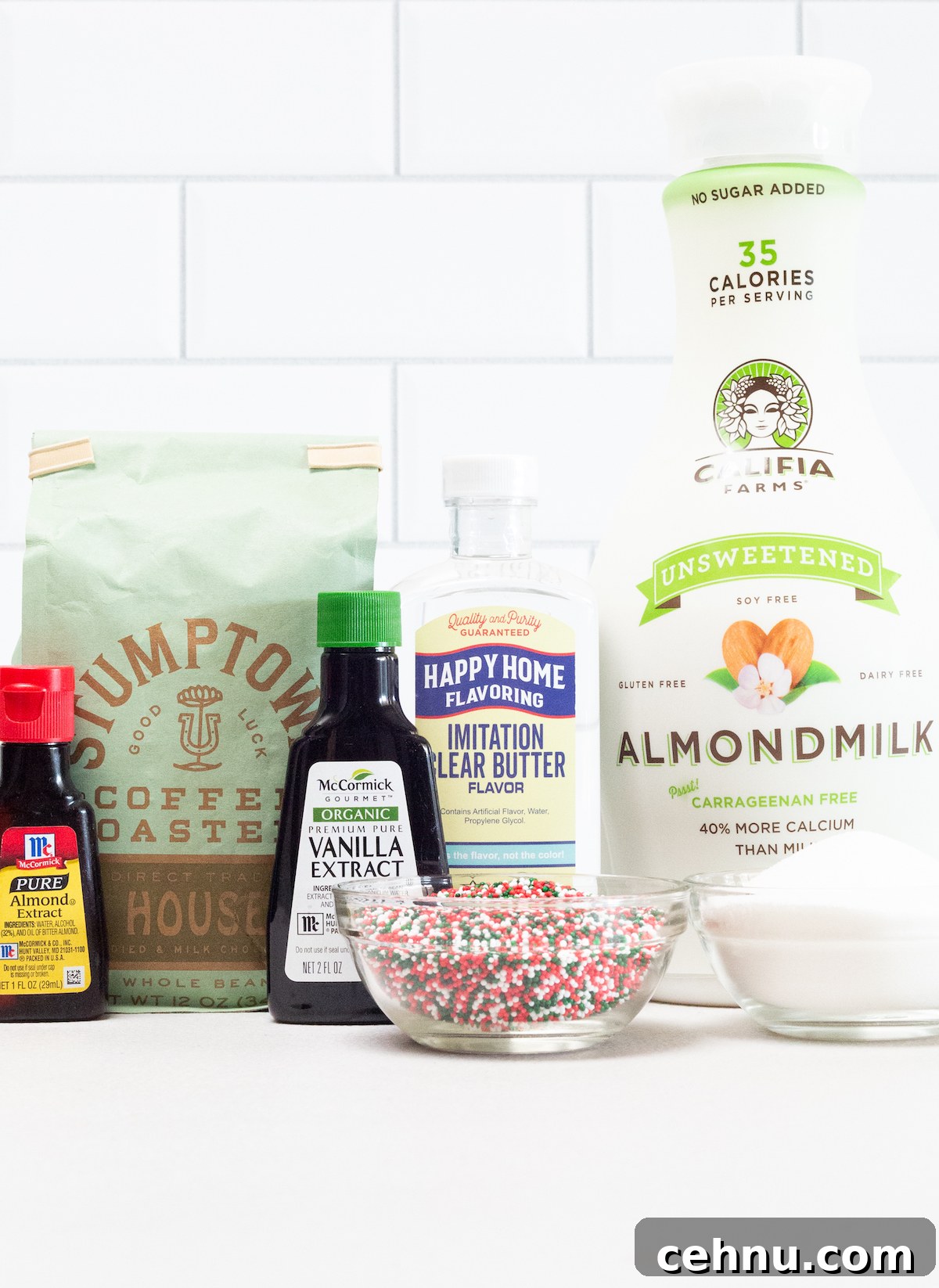 All the ingredients to make an iced sugar cookie latte on a white background. Almond extract, light roast coffee beans, vanilla extract, clear butter extract, almond milk, christmas sprinkles, and white granulated sugar.