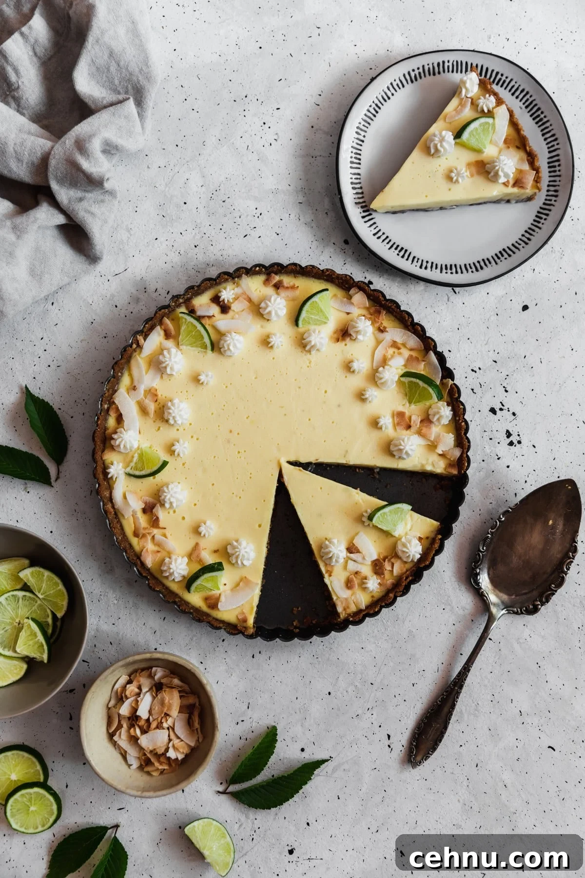Toasted Coconut Key Lime Tart Recipe 2 An overhead image of a sliced coconut key lime tart in a black pan on a grey table surrounded by a white plate topped with a tart slice, a beige linen, a vintage pie server, and a grey bowl of limes.