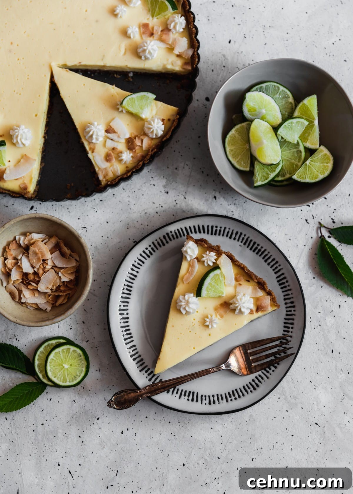 Toasted Coconut Key Lime Tart Recipe 3 An overhead image of a white plate with a slice of lime pie on a grey counter next to a whole pie, grey bowl of limes, and a small bowl of toasted coconut.