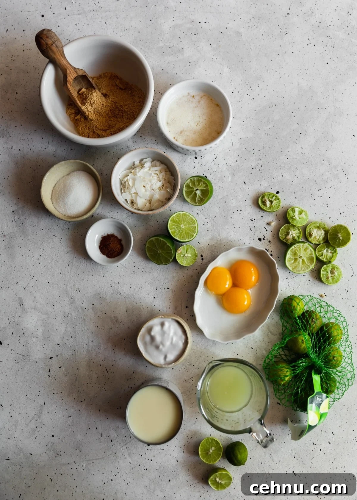 Toasted Coconut Key Lime Tart Recipe 4 An overhead image of various white and cream-colored bowls filled with ingredients for key lime pie on a grey speckled table next to a can of condensed milk and juiced key limes.