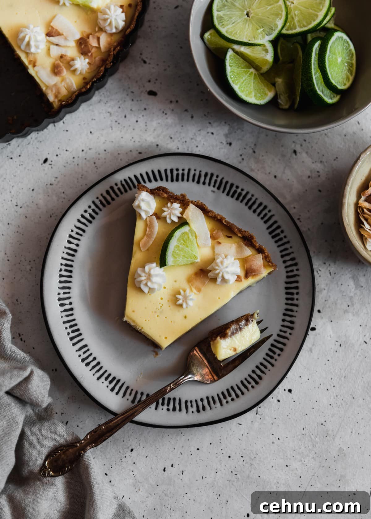 Toasted Coconut Key Lime Tart Recipe 6 A closeup overhead image of a white plate topped with a slice of coconut key lime pie with a bite taken out of it on a grey table next to a cream bowl of toasted coconut and grey bowl of limes.