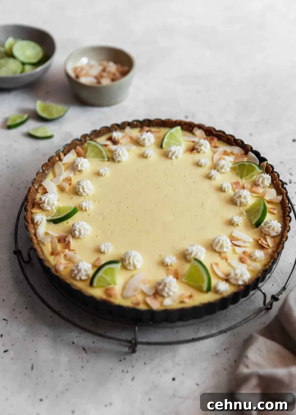 Toasted Coconut Key Lime Tart Recipe 7 A side image of a coconut key lime pie on a cooling rack placed on a grey table next to a bowl of coconut flakes, bowl of limes, and grey linen.