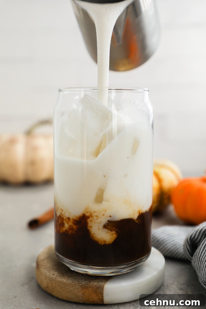 A glass filled with ice and pumpkin spice espresso is being filled with frothy milk and surrounded by small pumpkins by small pumpkins