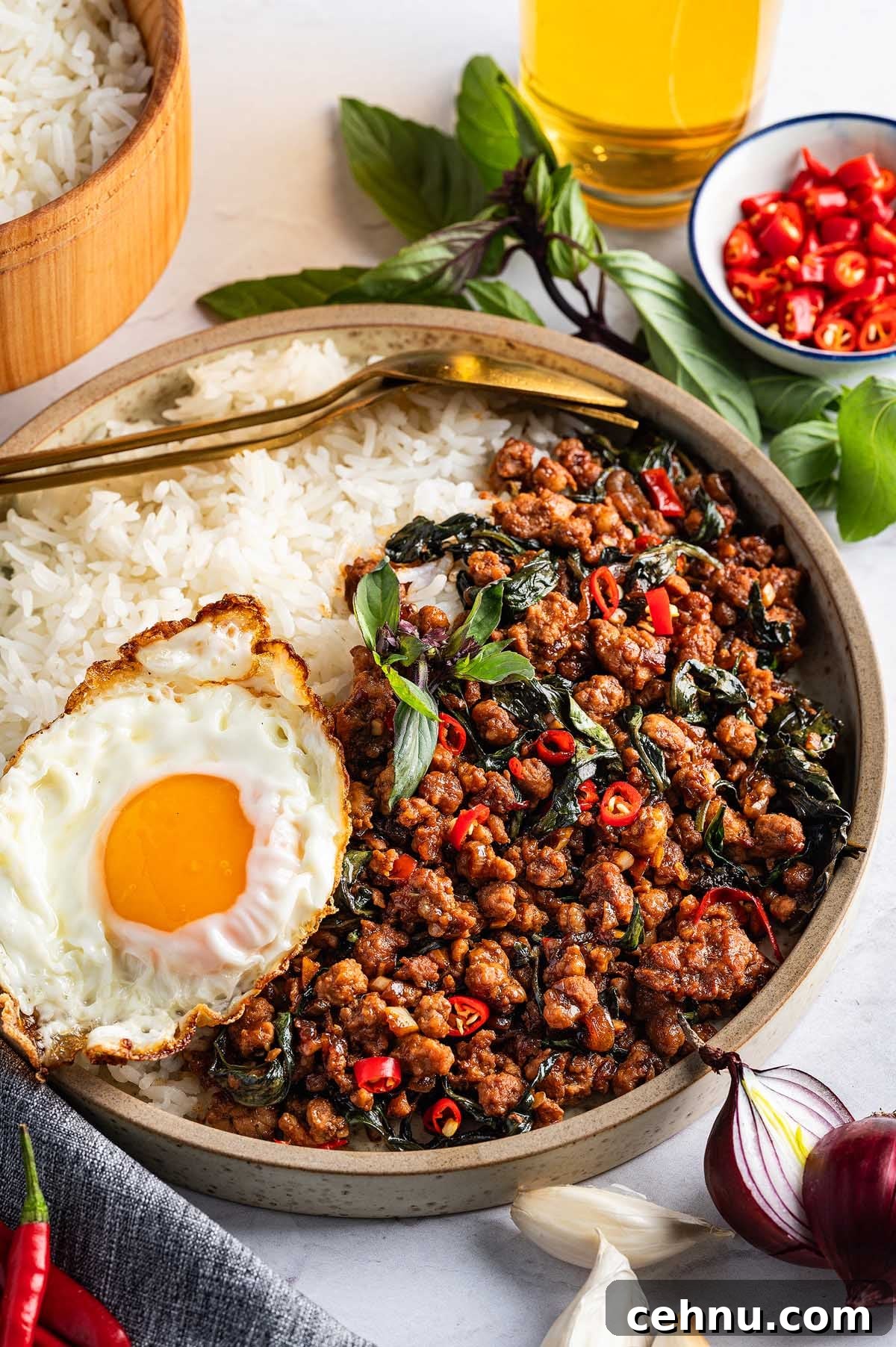 A plate of Thai basil pork, topped with minced meat and red chilies, is served alongside a fried egg. A glass of yellow beverage and a bowl of sliced red chilies add extra flavor to the meal.