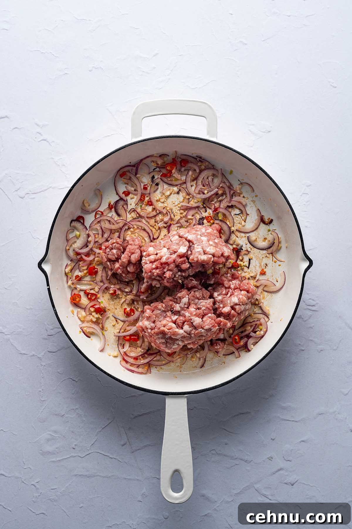 Ground meat with sliced onions, red chili peppers, and aromatic Thai basil pork sizzle in a white skillet on a gray surface.