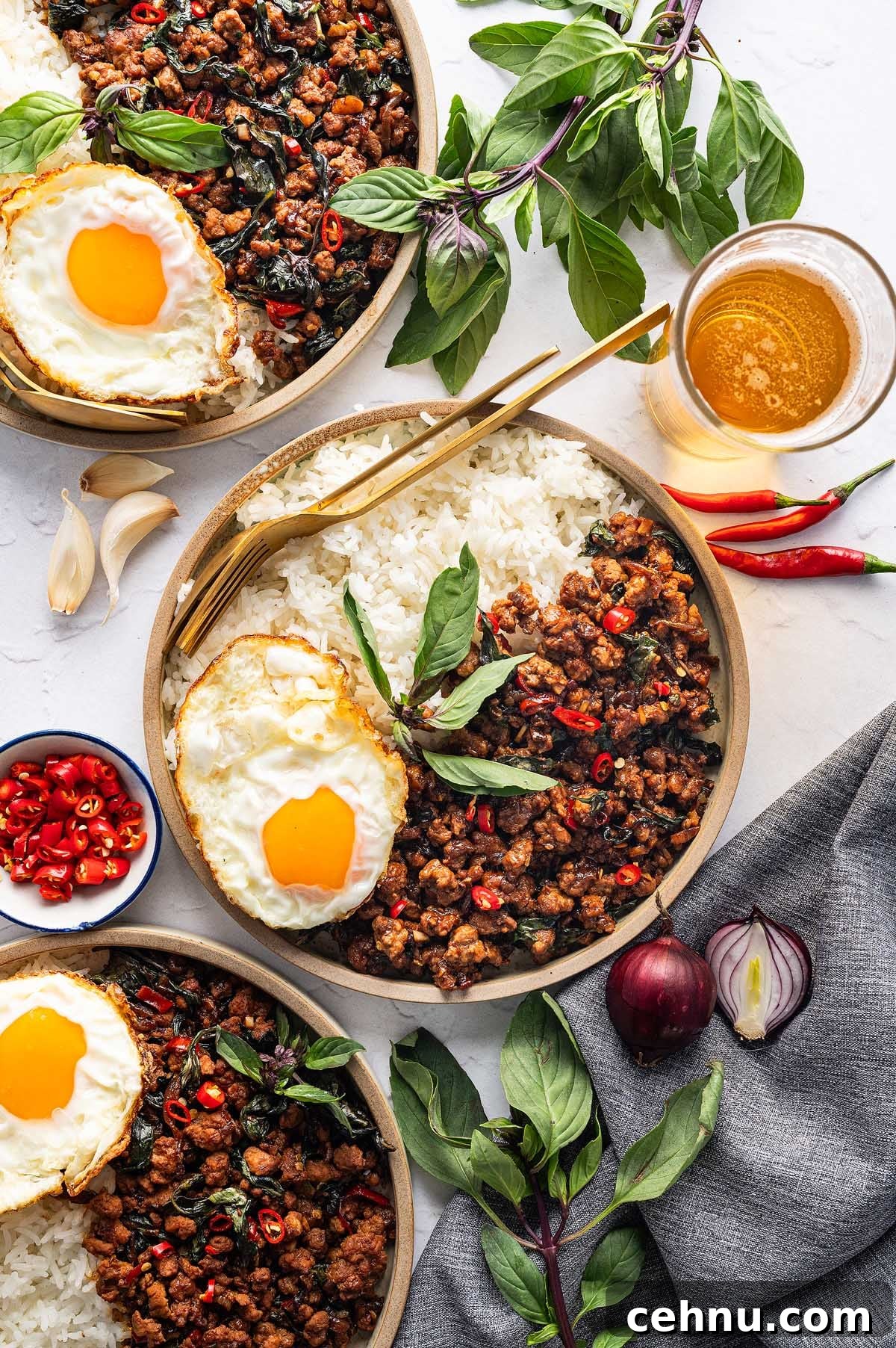 Three plates of rice topped with Thai basil pork and a fried egg each, surrounded by fresh basil, chili peppers, garlic, and accompanied by a glass of beer on a light surface.