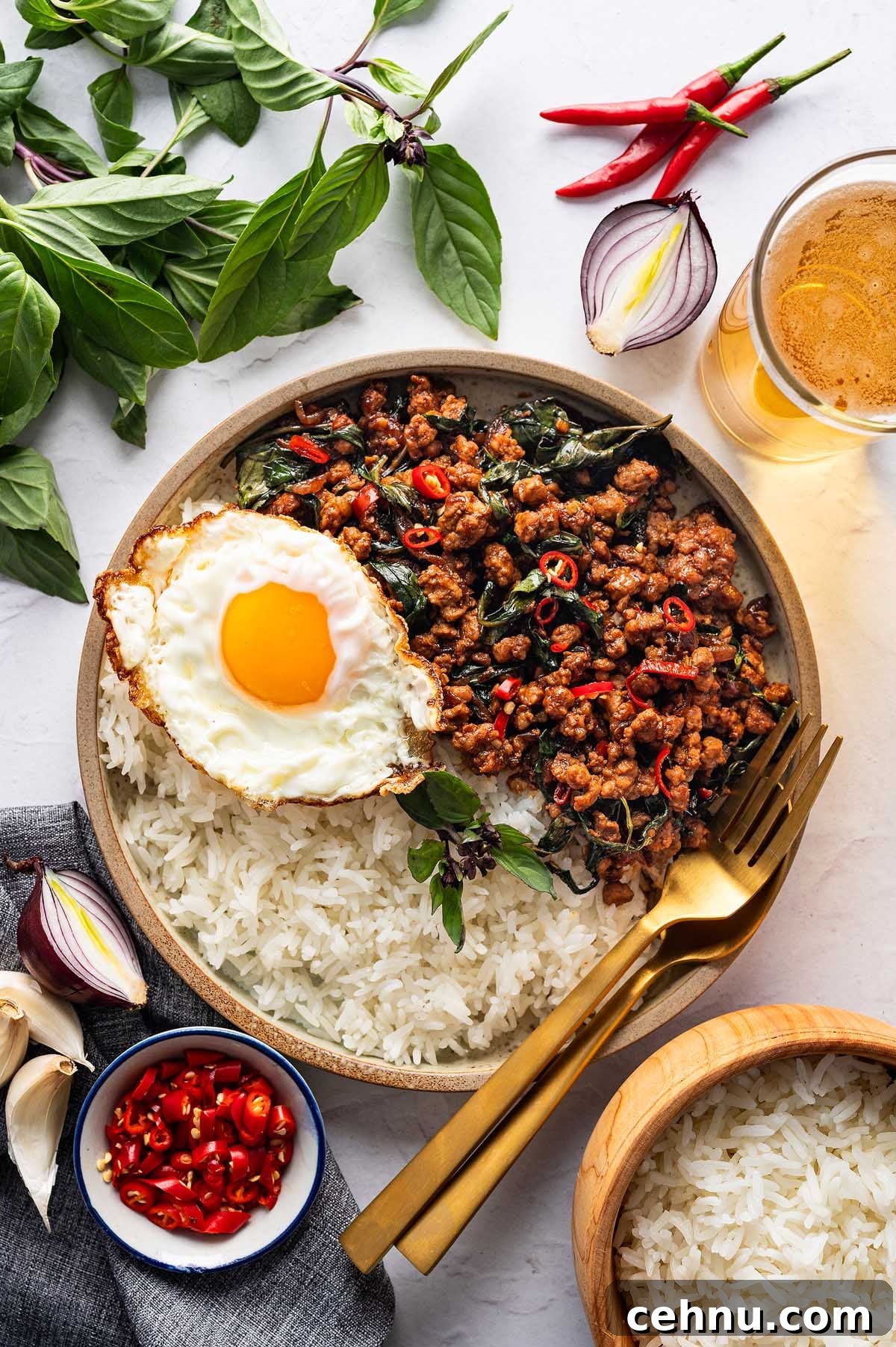 A plate of rice topped with Thai basil pork, stir-fried to perfection with chili and basil, accompanied by a fried egg. Garnished with red chilies and fresh basil leaves, and complemented by a refreshing drink nearby.