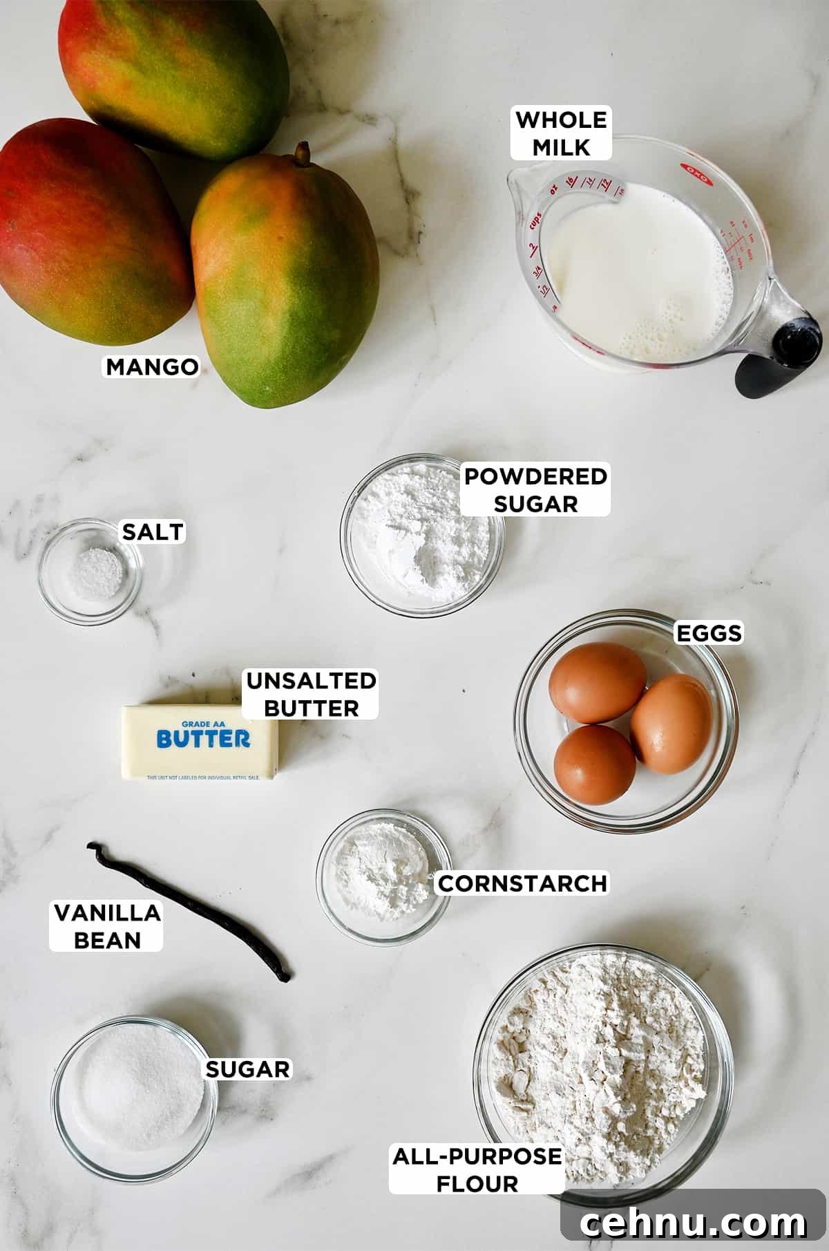 Three ripe mangoes next to a liquid measuring cup filled with whole milk and various sizes of glass bowls containing powdered sugar, eggs, a stick of unsalted butter, salt, a vanilla bean, flour and cornstarch.