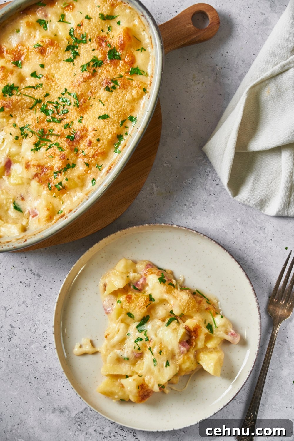 Ham and potato casserole on a white plate with a fork next to the plate and the rest of the casserole on top of a wooden board.