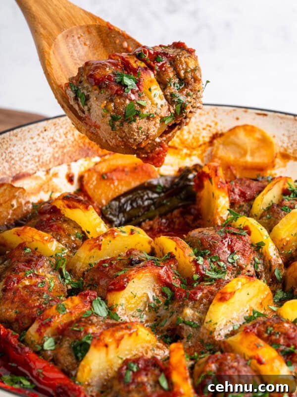 A wooden spoon holds a seasoned meatball from a dish with sliced potatoes, herbs, and bell peppers, all baked in a tomato sauce.