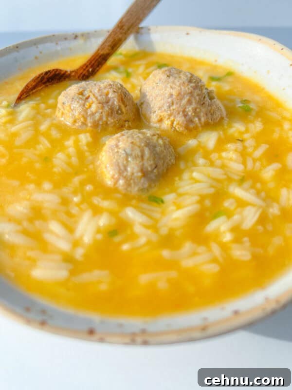 A bowl of Hidden Veggie Meatball Soup with yellow broth, rice, and three meatballs, served with a wooden spoon resting on the side.