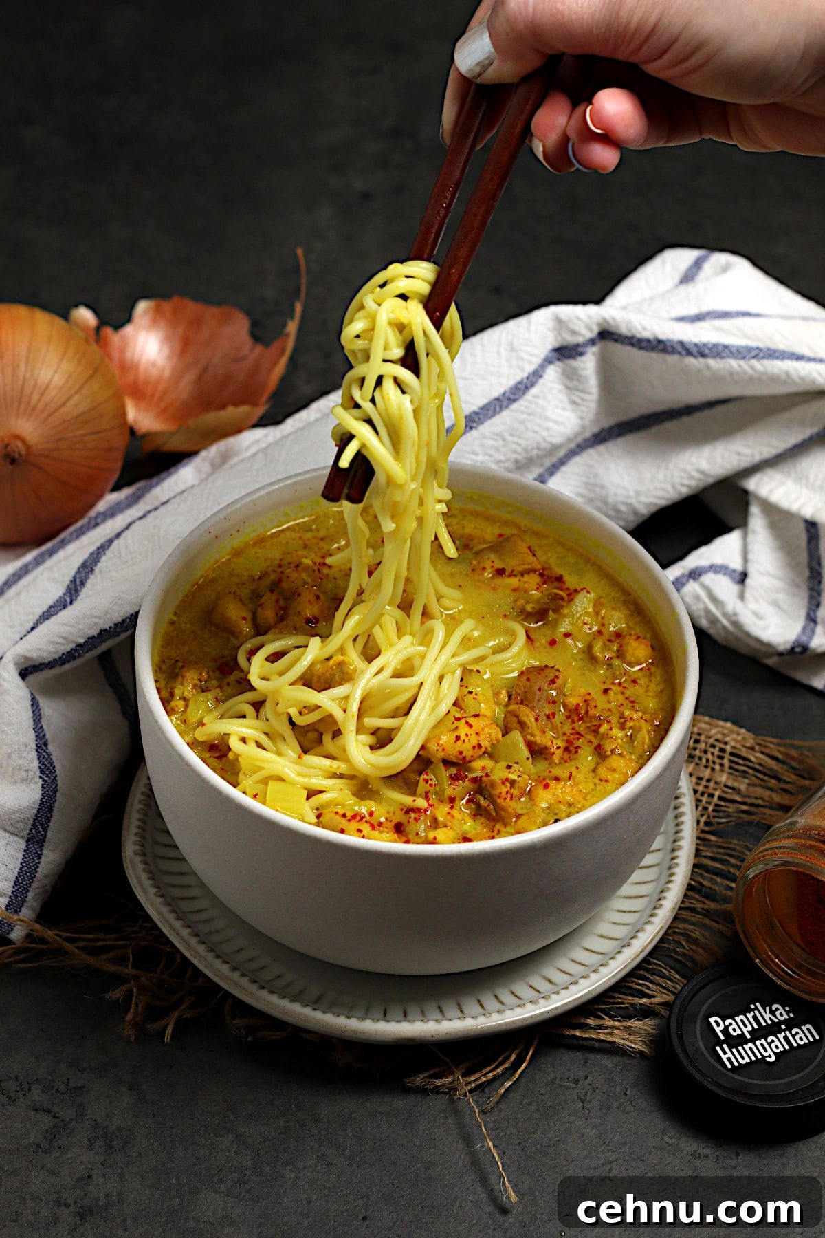 A hand pulling noodles from a bowl of Burmese Coconut Chicken Noodle soup with wooden chopsticks.
