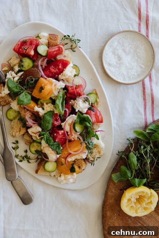 panzanella salad on a white oval platter on top on a linen tablecloth with a salt pinch pot in the top right and fresh herbs and lemon in the bottom right.