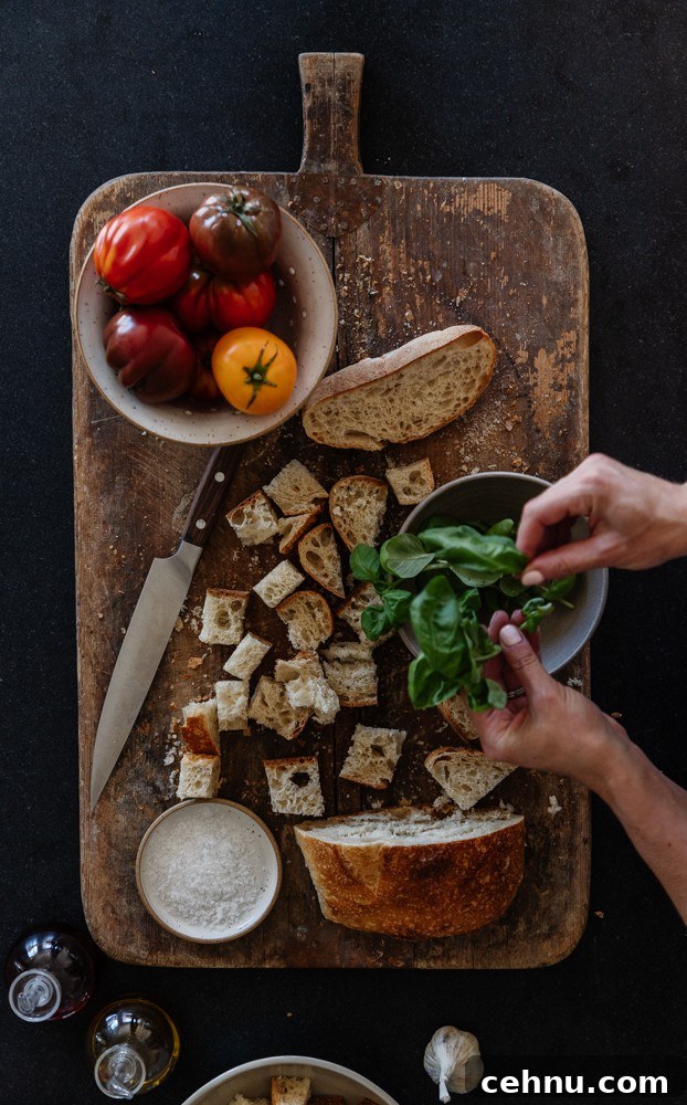 rustic cutting board on black granite with cubed sourdough, bowl of tomatoes, and hands tearing some fresh basil