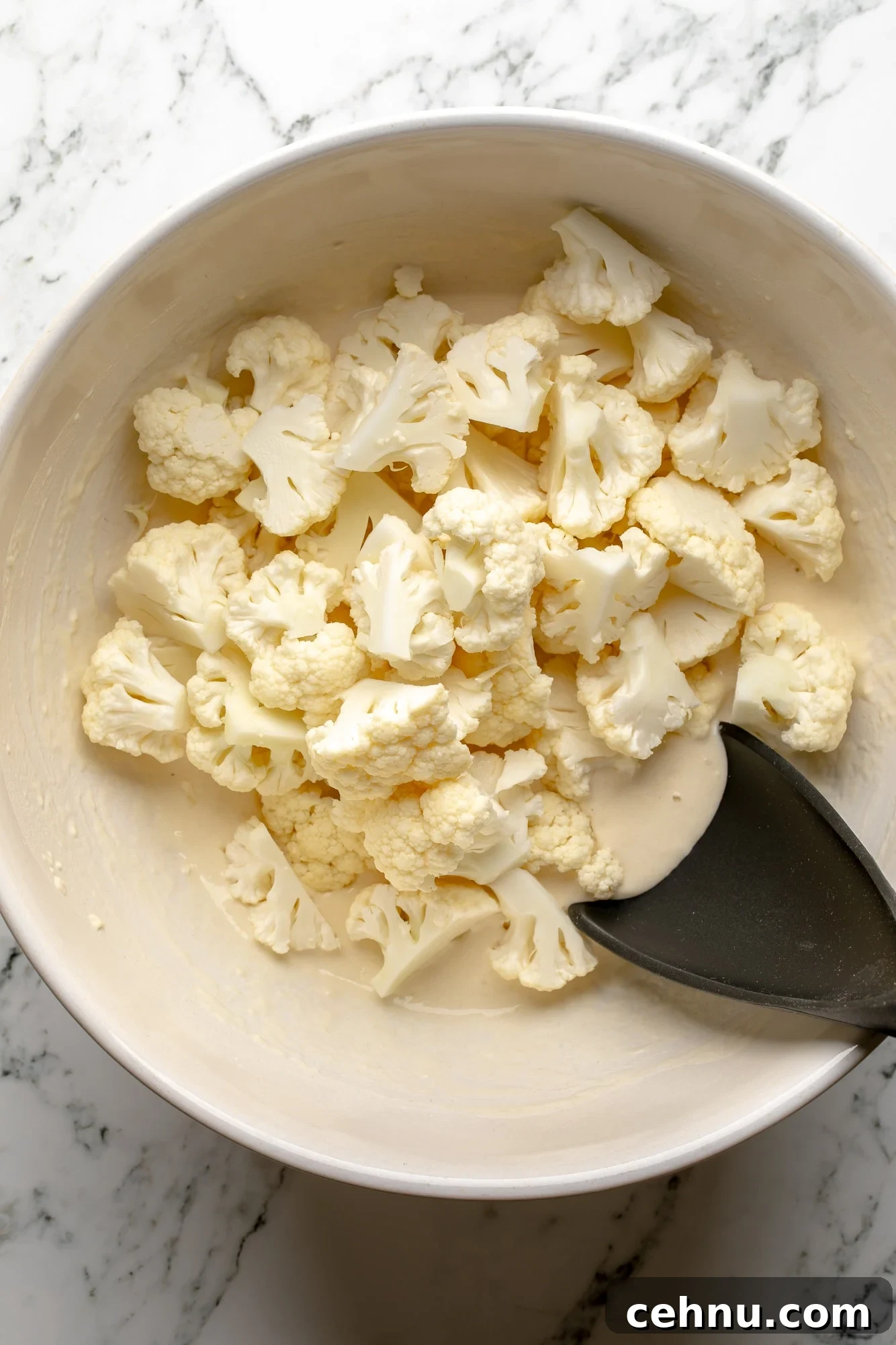 Overhead view of cauliflower florets being tossed in a batter in a large white bowl.