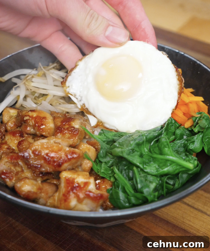 Fried egg being placed on top of a chicken bibimbap bowl with chicken, vegetables, and rice before adding sauce.