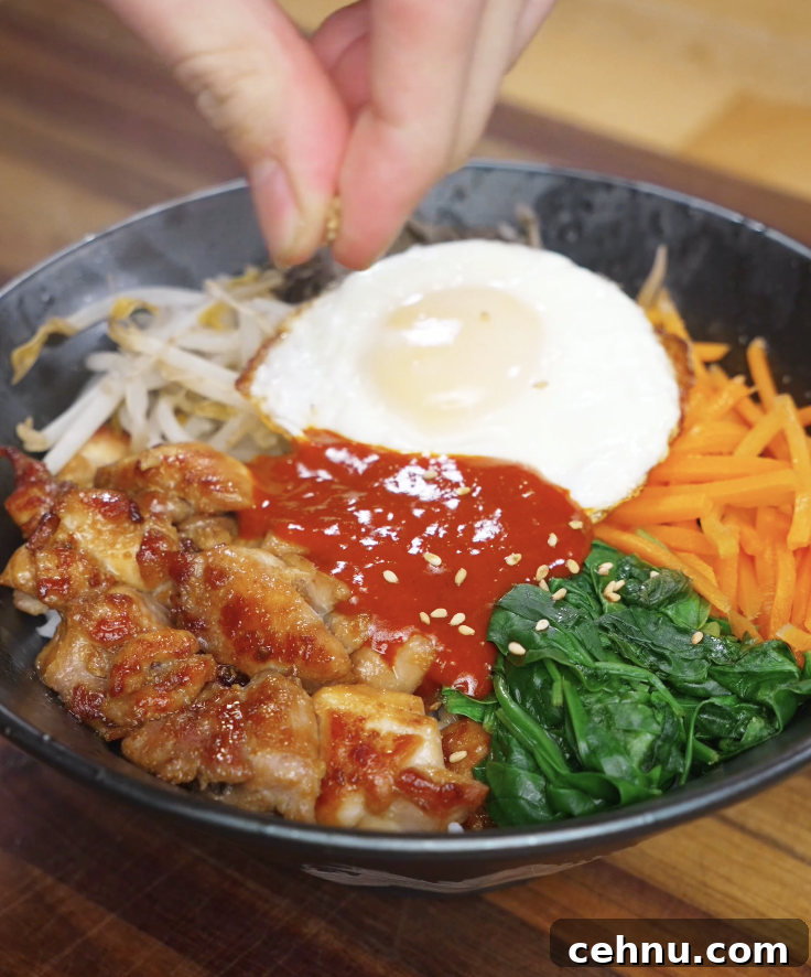 Sprinkling sesame seeds over chicken bibimbap with gochujang sauce, fried egg, and vegetables as the final garnish step.