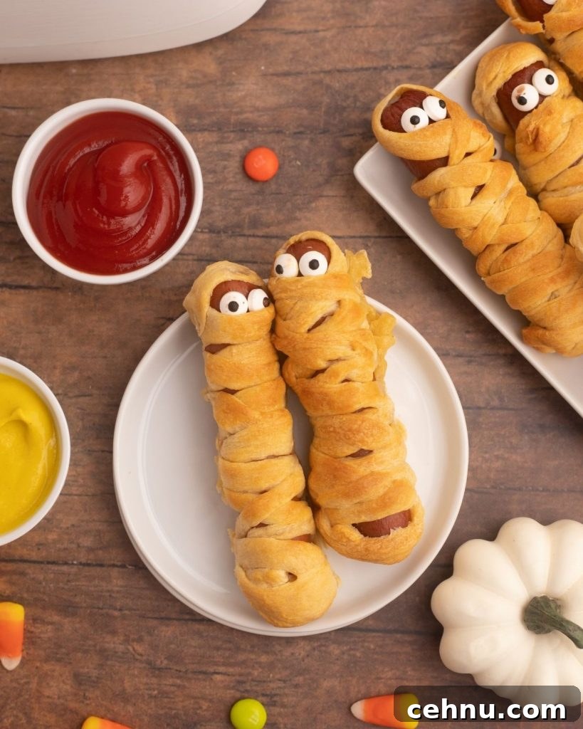 Air Fryer Mummy Hot Dogs: Halloween Appetizer Recipe 3 Golden hot dog mummies on a white plate with mustard and ketchup and candies scattered around the table.