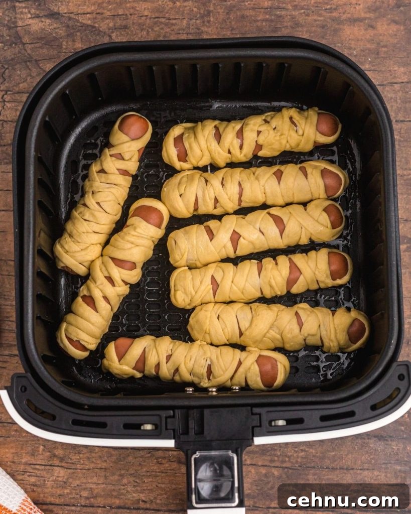 Air Fryer Mummy Hot Dogs: Halloween Appetizer Recipe 6 Hot dogs wrapped in crescent roll strips to look like mummies in the air fryer basket before being cooked.