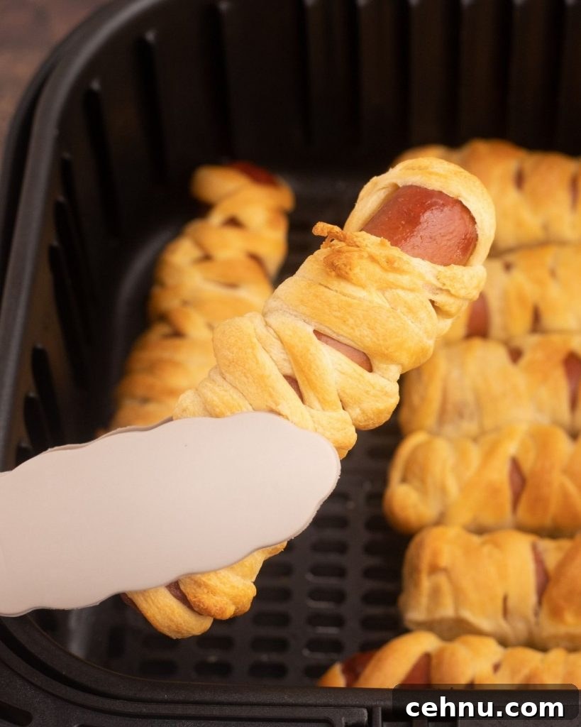 Air Fryer Mummy Hot Dogs: Halloween Appetizer Recipe 8 Crusted wrapped hot dog after being cooked until golden and being lifted out of basket with tongs.