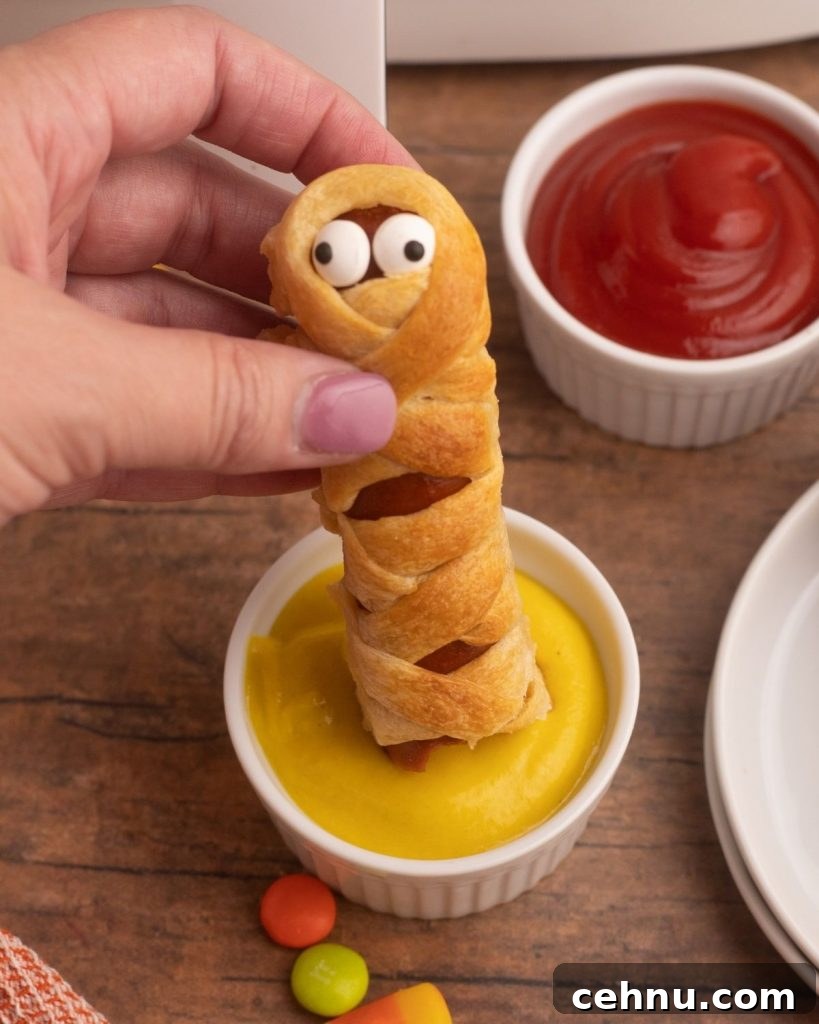 Air Fryer Mummy Hot Dogs: Halloween Appetizer Recipe 9 Close up photo of crust wrapped hot dog being dipped into mustard with candies scattered on a wooden table.