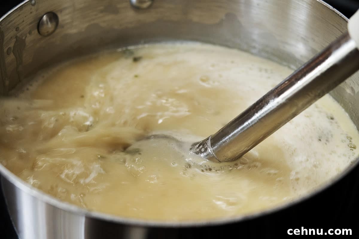 Creamy White Bean Soup with Garlic and Herbs 13 A metal pot on a stove with creamy white bean soup being pureed by an immersion blender. The soup is creamy and frothy, with visible steam rising from the surface.