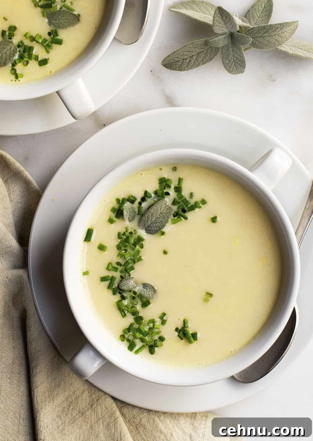 Creamy White Bean Soup with Garlic and Herbs 4 A white bowl of creamy white bean soup garnished with chopped chives and sage leaves. The bowl is on a saucer with a spoon beside it, and a napkin is nearby. Another bowl is partially visible in the background on a marble surface.
