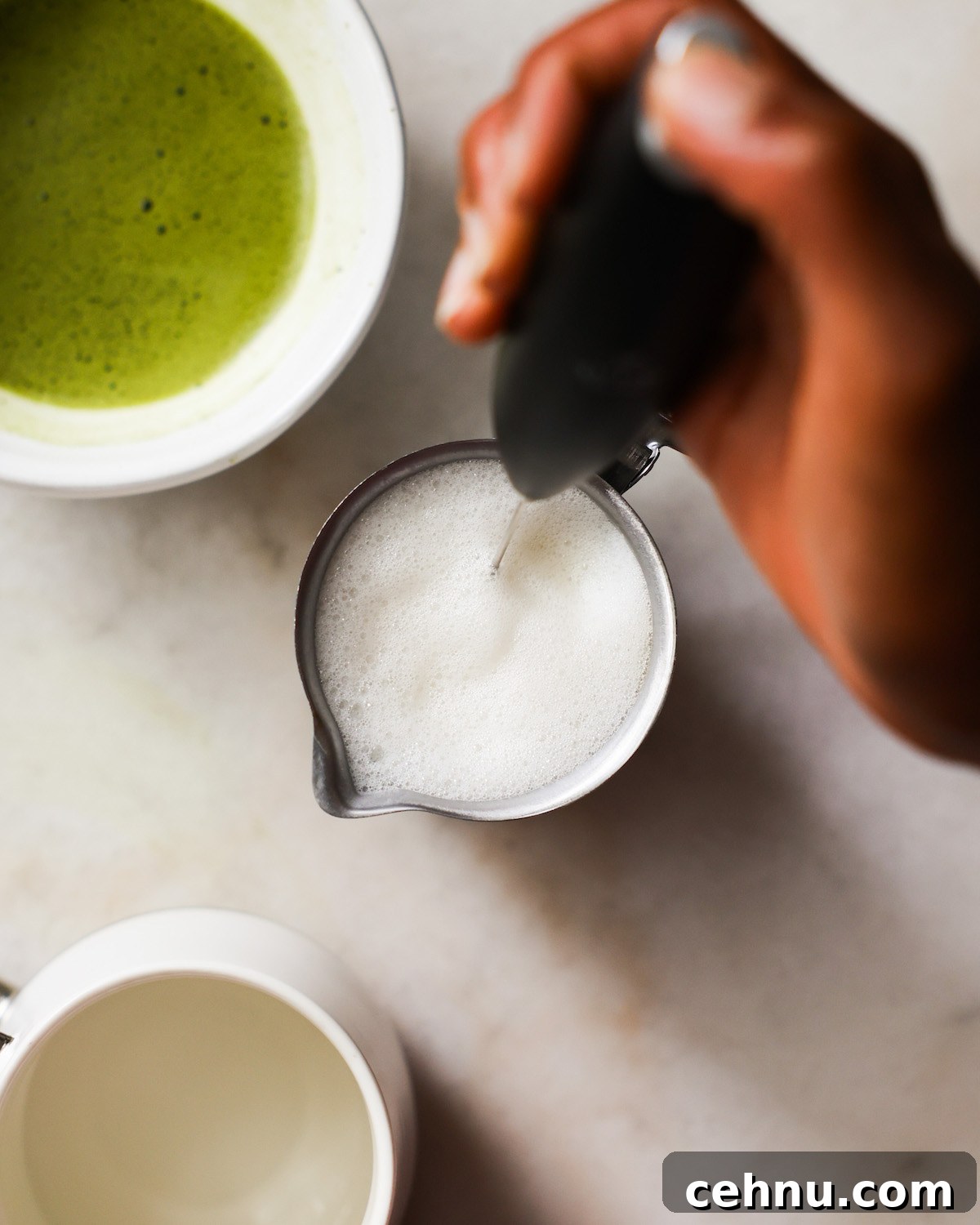 A brown hand frothing a small pitcher of oat milk.