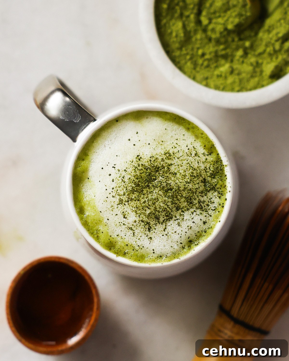 An overhead shot of a mug of the oat milk matcha latte on the counter next to a small dish of matcha and the matcha whisk.