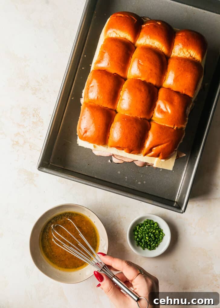 Oven-Baked Brown Sugar Ham and Melted Cheese Sliders 7 A hand whisking butter Dijon topping in a grey bowl next to a pan of sliders and a white bowl of chives on a tan counter.
