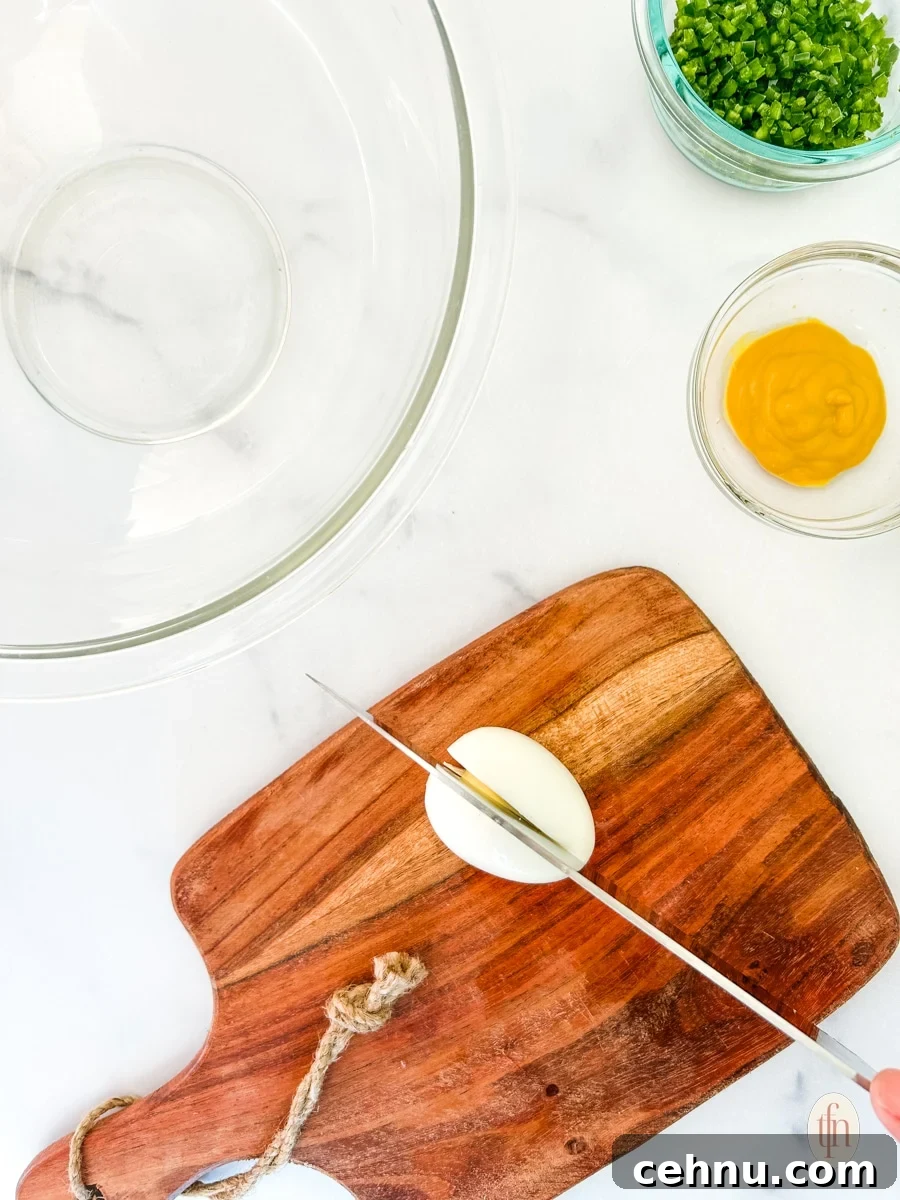 Egg being sliced on a wooden chopping board with a glass mixing bowl and a couple of prepared ingredients.