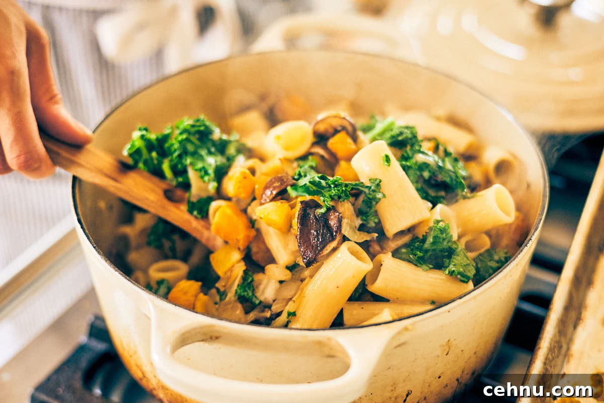 Roasted butternut squash, mushrooms, and kale being stirred into a pot of pasta.