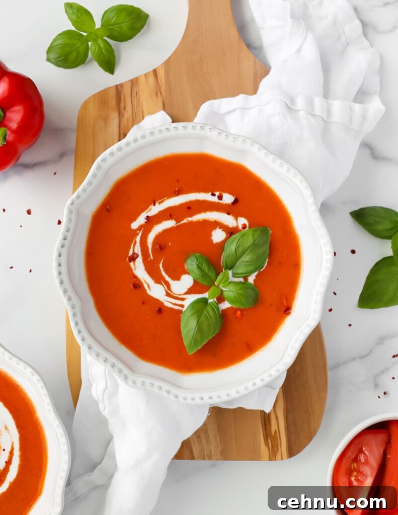 A white bowl filled with red soup that is garnished with white coconut milk, fresh green basil leaves, and crushed red pepper.