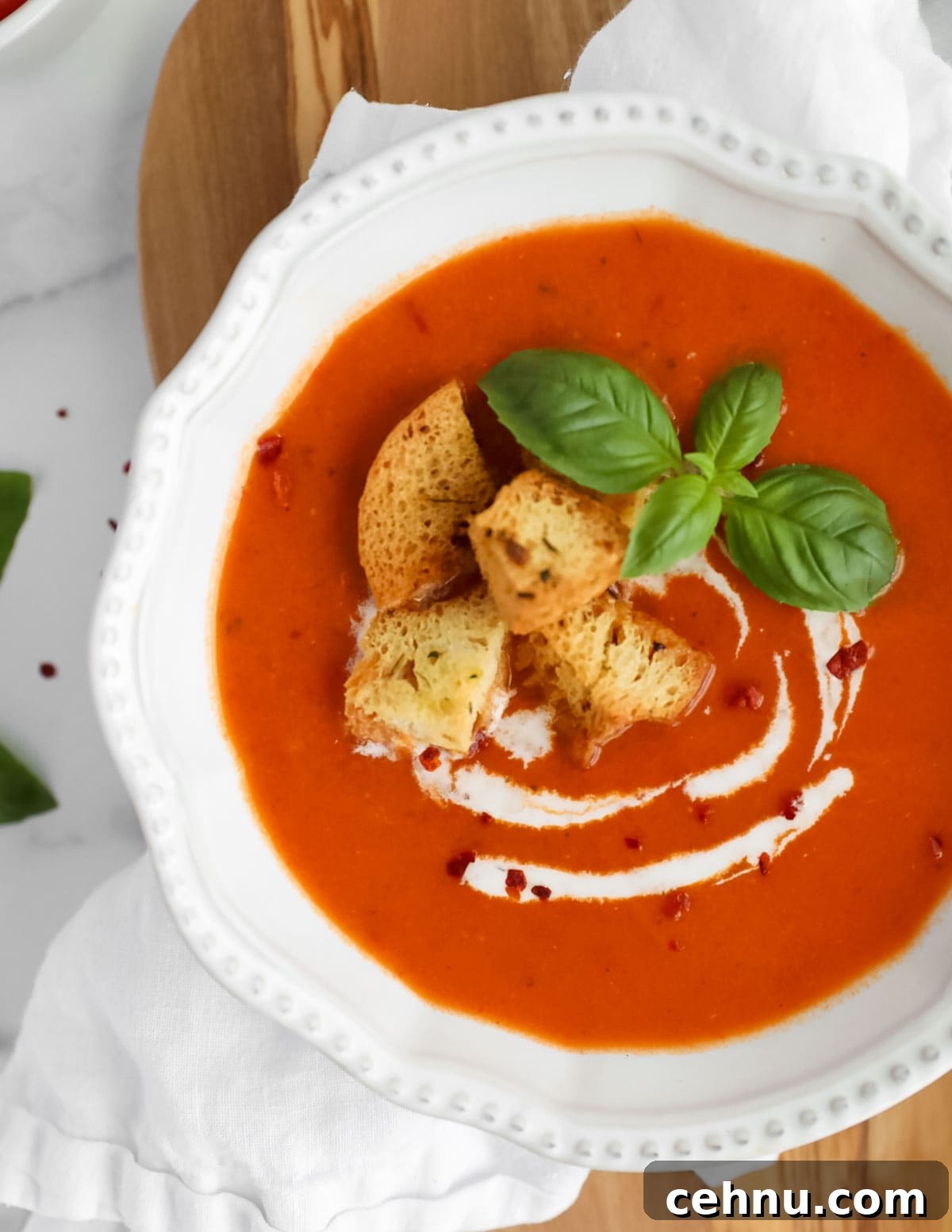 An overhead image of red soup in a white bowl garnished with croutons, basil, coconut milk, and crushed red pepper flakes.