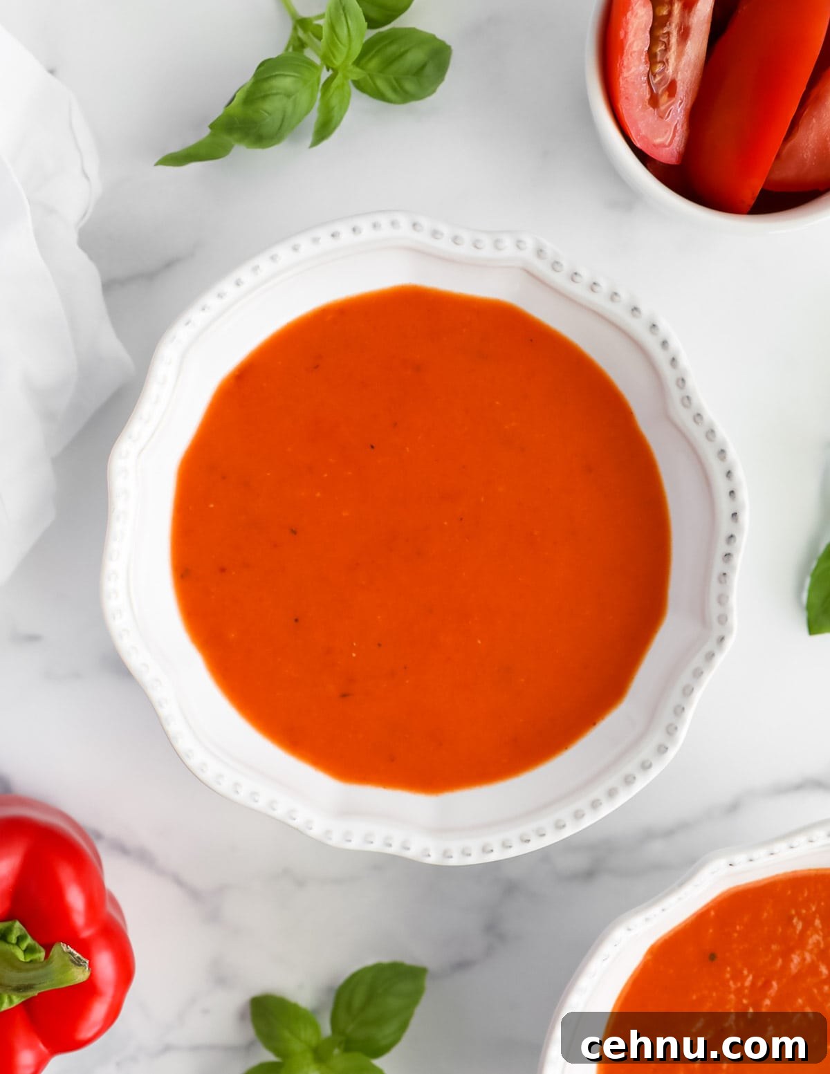 A white bowl filled with creamy red soup. In the background there is a red bell pepper, a bowl of sliced tomatoes, and leaves of basil