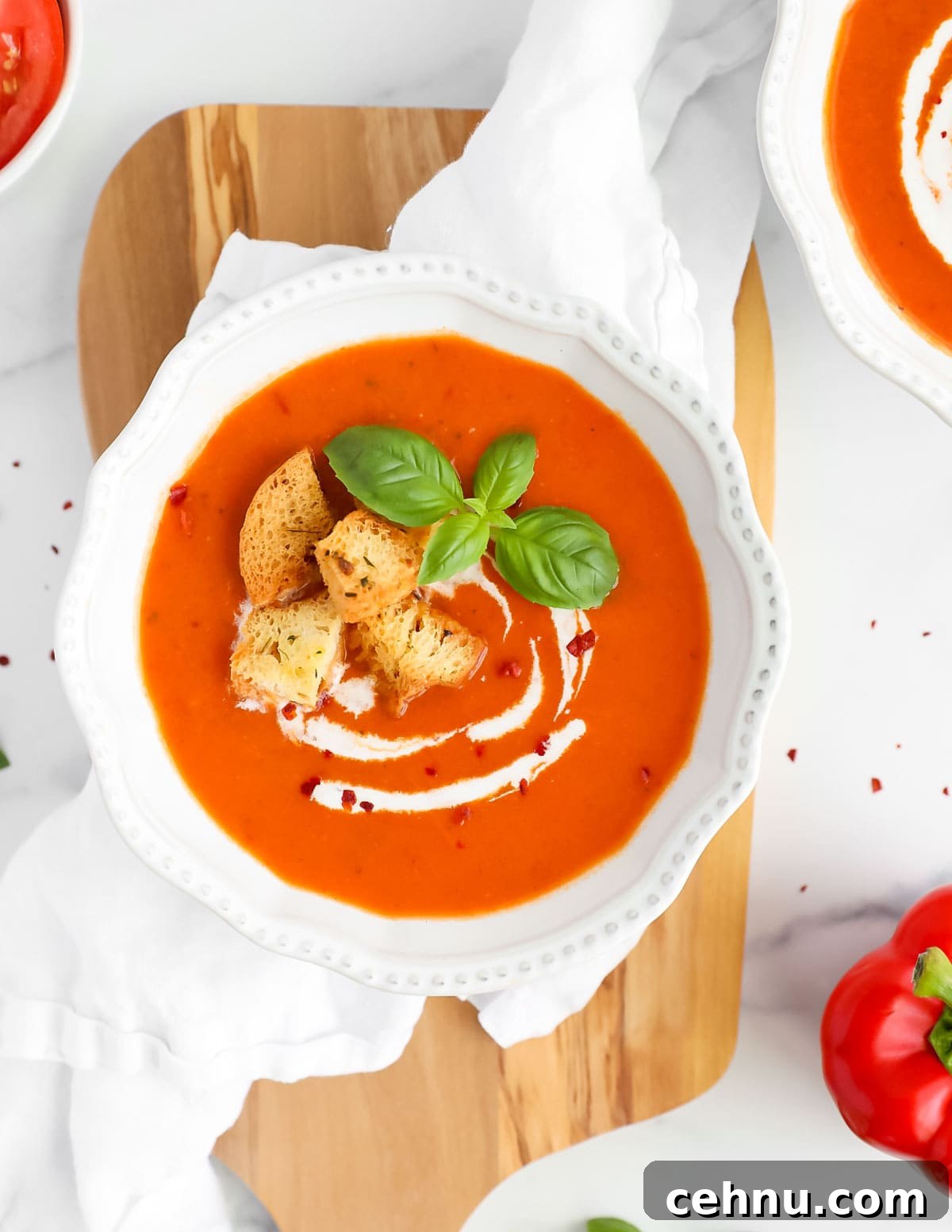 An overhead image of red soup in a white bowl garnished with coconut milk drizzle, croutons, basil leaves, and crushed red pepper