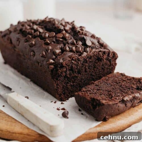 A chocolate loaf cake on parchment paper topped with chocolate chips, one slice has been cut.