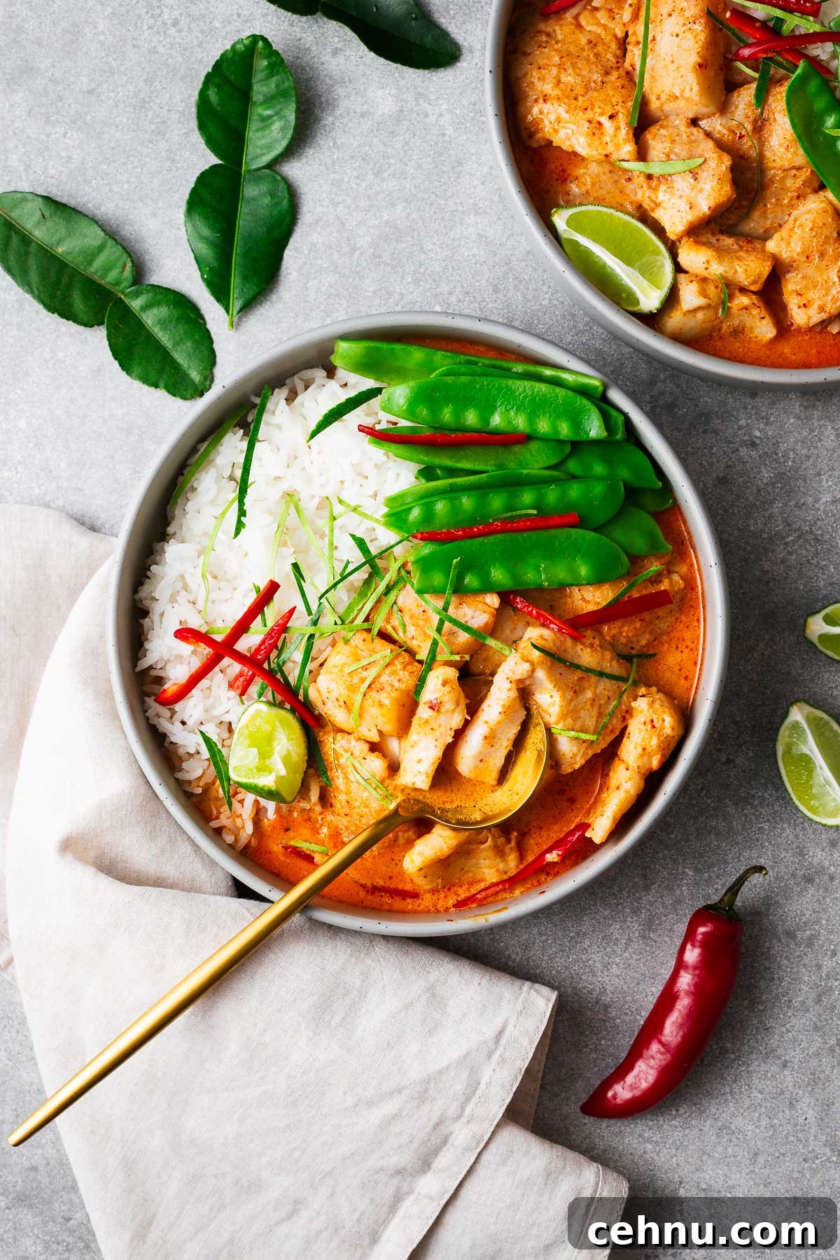 A bowl with steamed jasmine rice, Thai fish red curry and snow peas topped with sliced red chile and kaffir lime leaves wit a golden serving spoon.