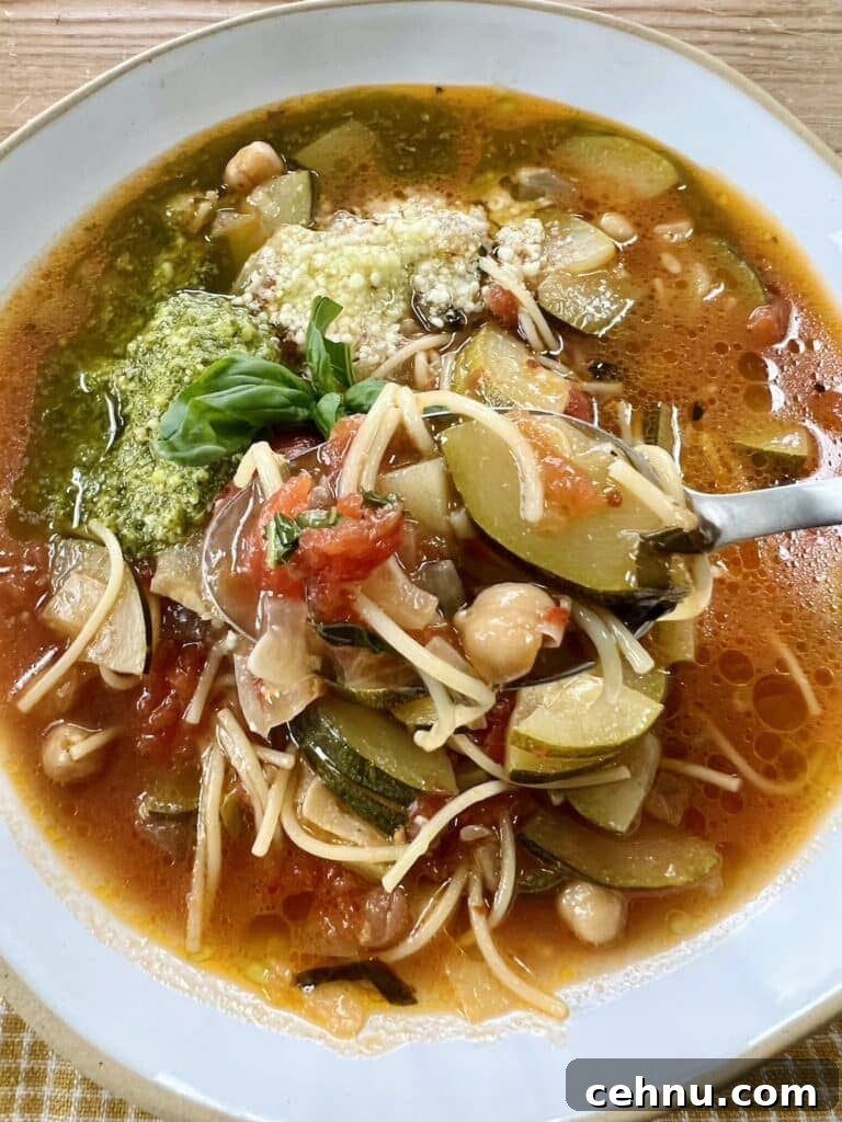 Italian zucchini soup in white bowl with basil pesto, grated cheese and basil sprig garnish with spoon in bowl.