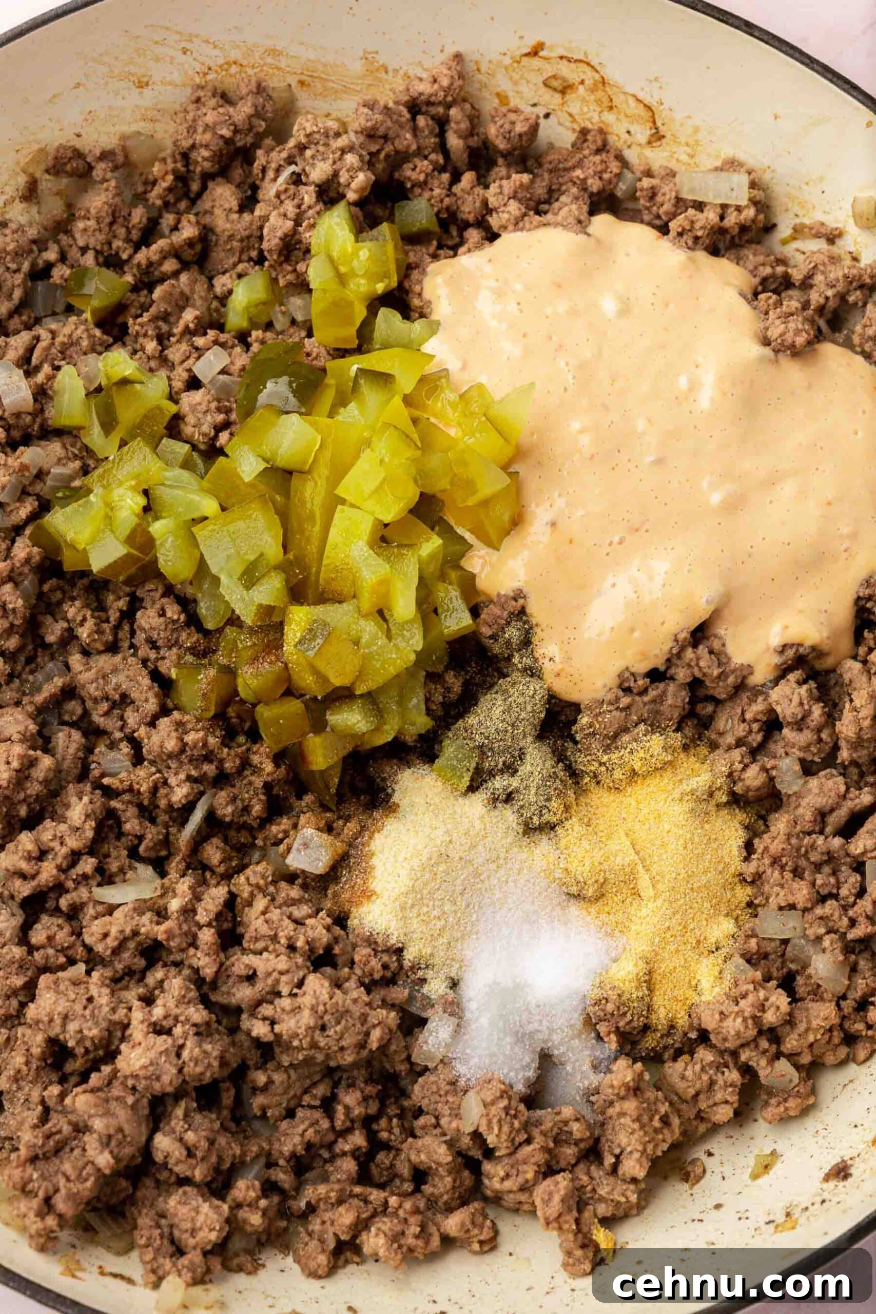 Cooked ground beef in a braising pan topped with diced pickles, garlic powder, salt, onion powder and thousand island dressing before mixing together.