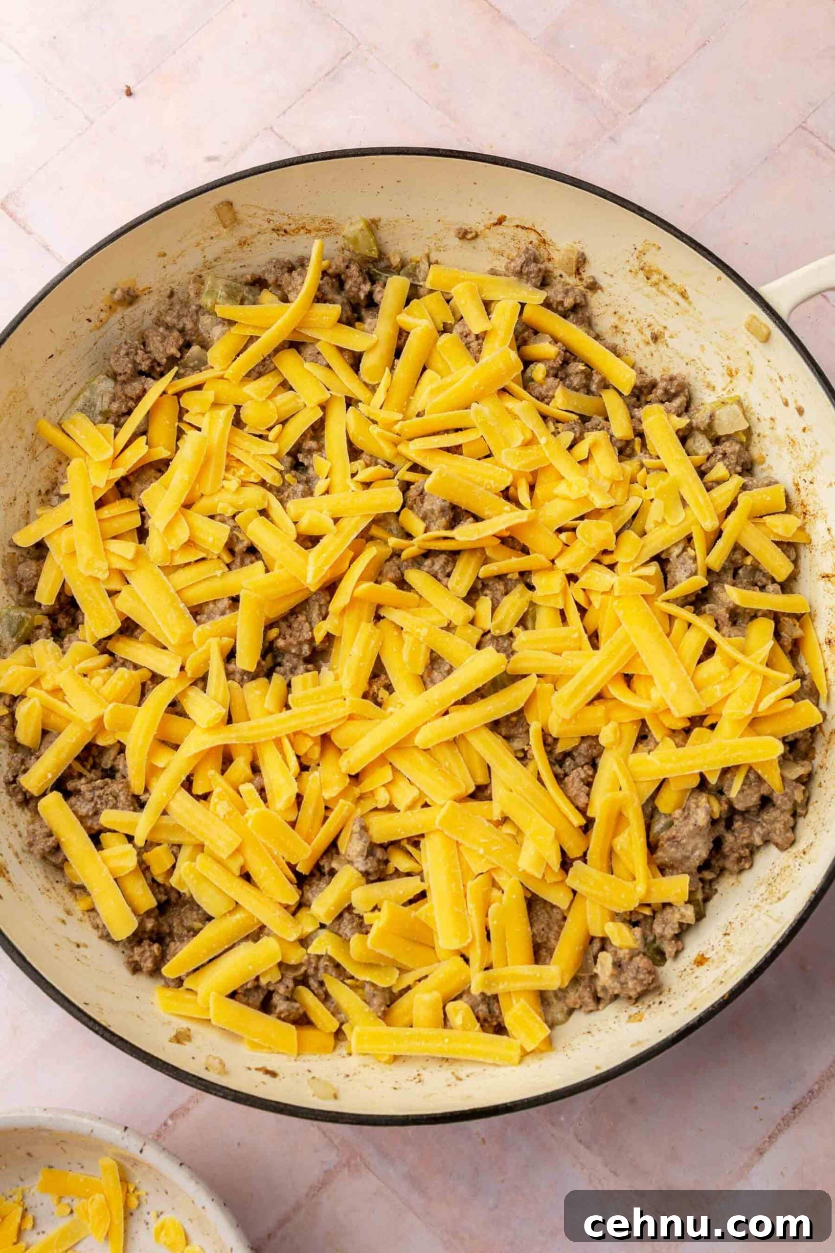 A braising pan with a ground beef mixture topped with shredded cheddar cheese before baking in the oven.