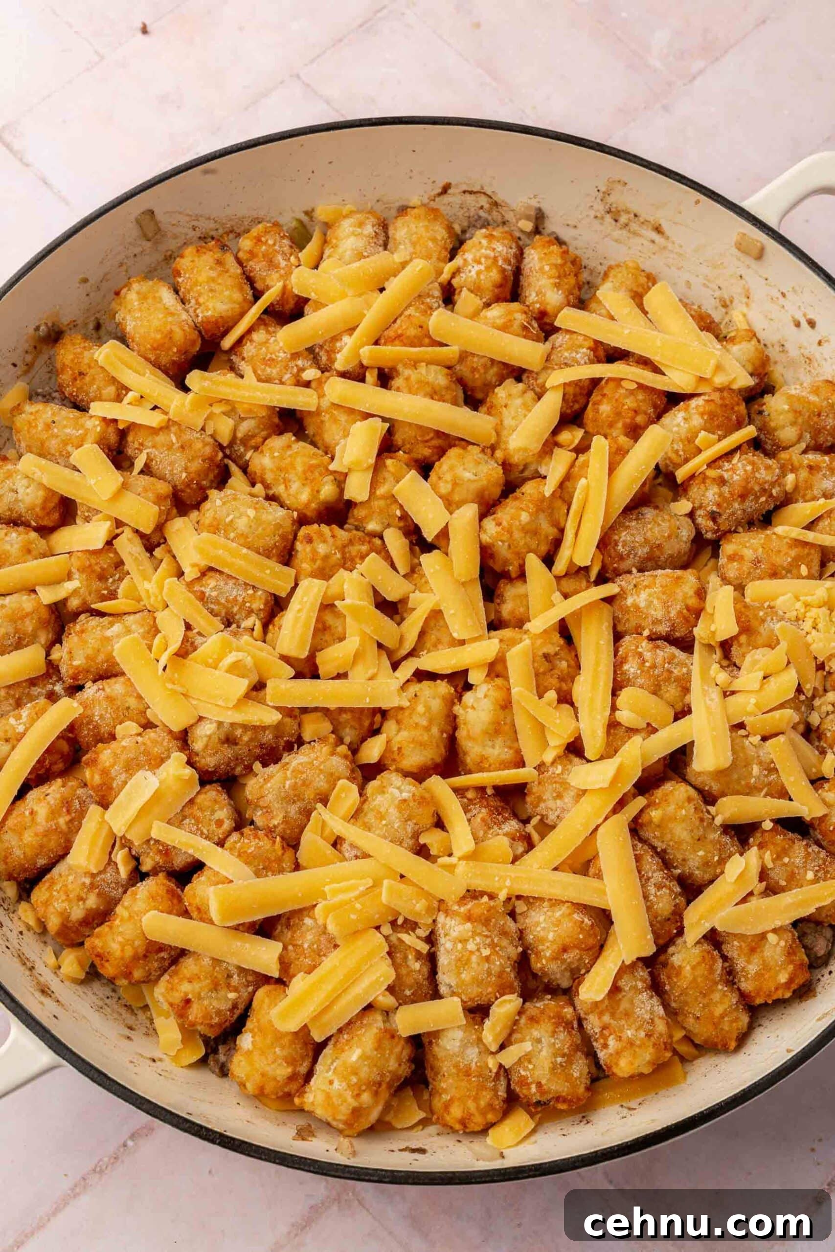 Tater tots topped with shredded cheese in a round white braising pan before placing in the oven.